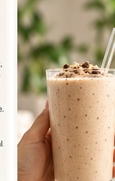 Hand holding a tall creamy shake topped with cookie pieces beside soft-focus greenery.