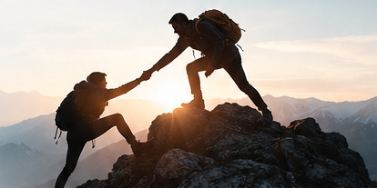 One hiker reaching down to help another climb a rocky summit at sunset